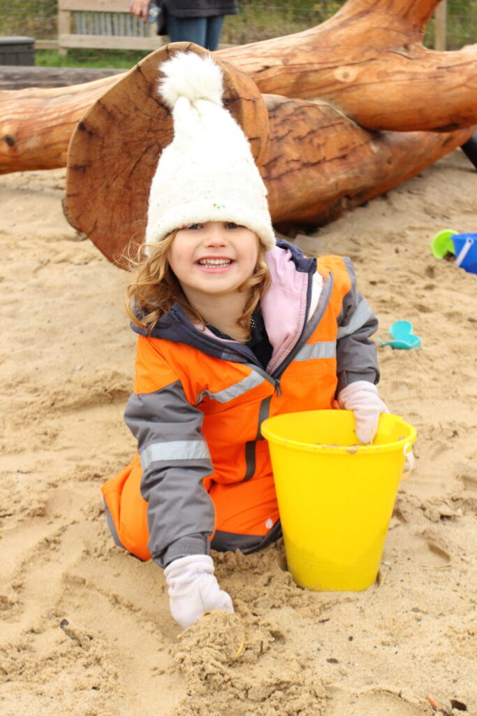 Child playing in sand with a yellow bucket, wearing cozy hat and colorful jacket.