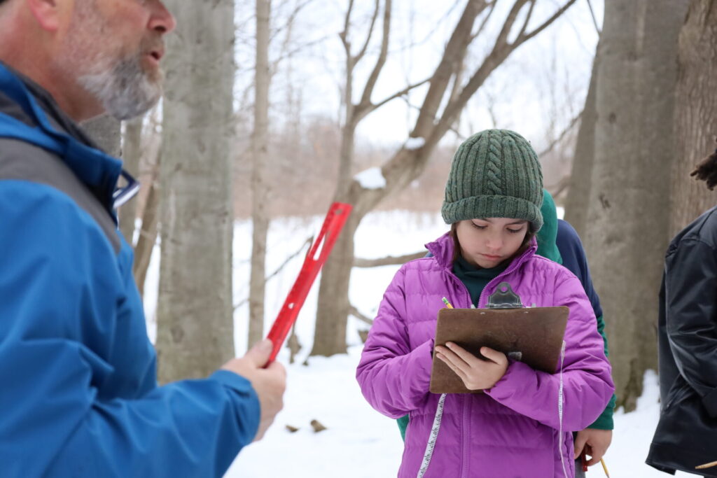 Young girl in snow checks clipboard during winter outdoor education with adults and friends.